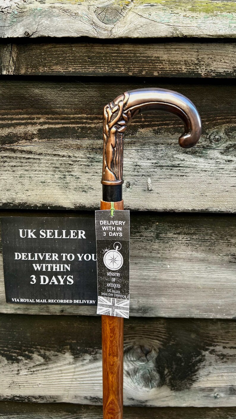 May include: A wooden walking stick with a copper-colored handle featuring a detailed design. The handle is curved and has a decorative design. The stick is brown and has a natural wood grain. The stick is accompanied by a tag that reads "Delivery within 3 days" and "Ministry of Antiques UK Seller Same Day Dispatch".