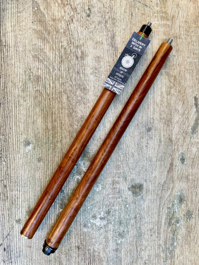 May include: Two brown wooden walking sticks with black metal tips. The sticks are on a wooden surface. A tag is attached to one stick that says "Delivery within 3 days" and "Ministry of Antiques, Established 1942, London, England".