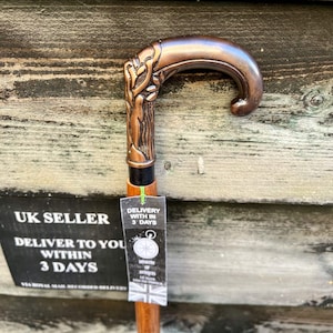 May include: A copper-colored walking stick with a detailed, ornate handle. The handle is shaped like a curved branch with a leaf design. The stick is made of wood and has a black shaft.