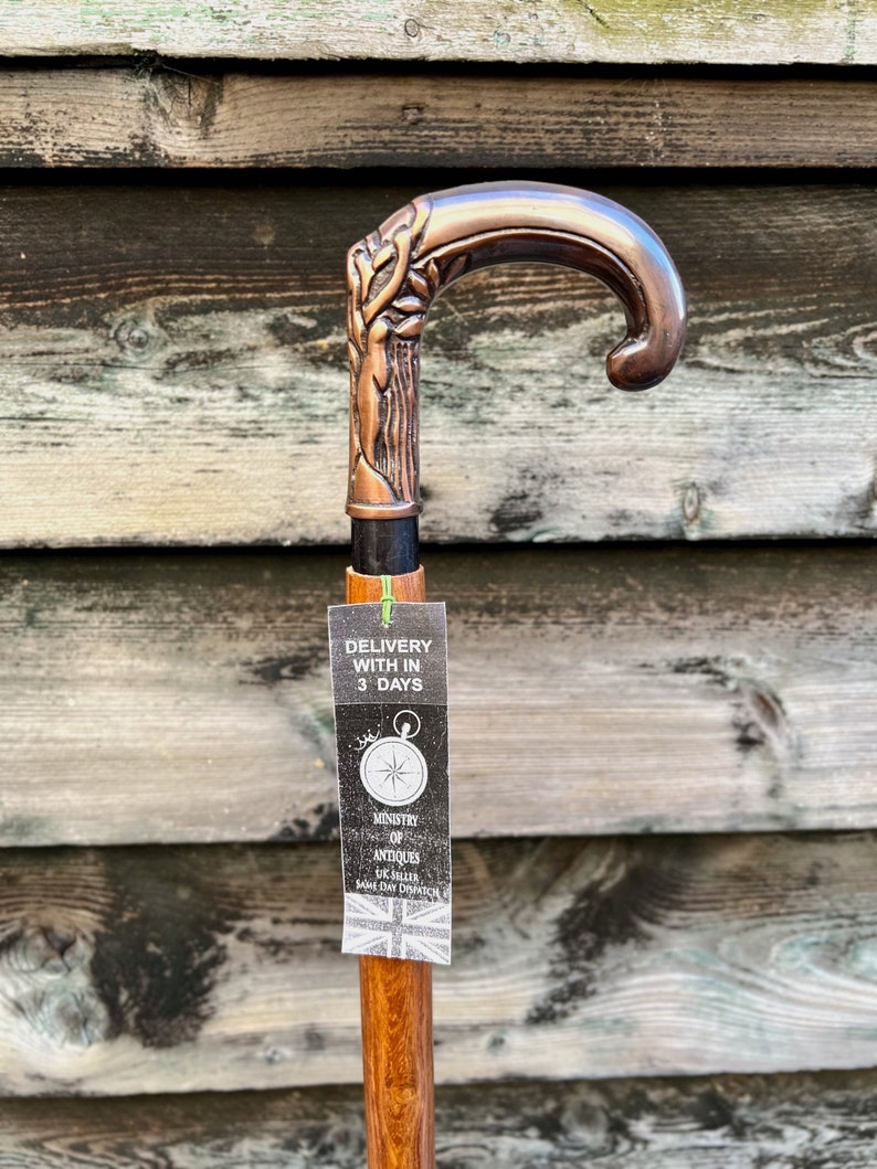 May include: A copper-colored walking stick with a detailed tree design on the handle. The stick has a wooden shaft and a tag that says "Delivery with in 3 days" and "Ministry of Antiques UK Seller Same Day Dispatch".