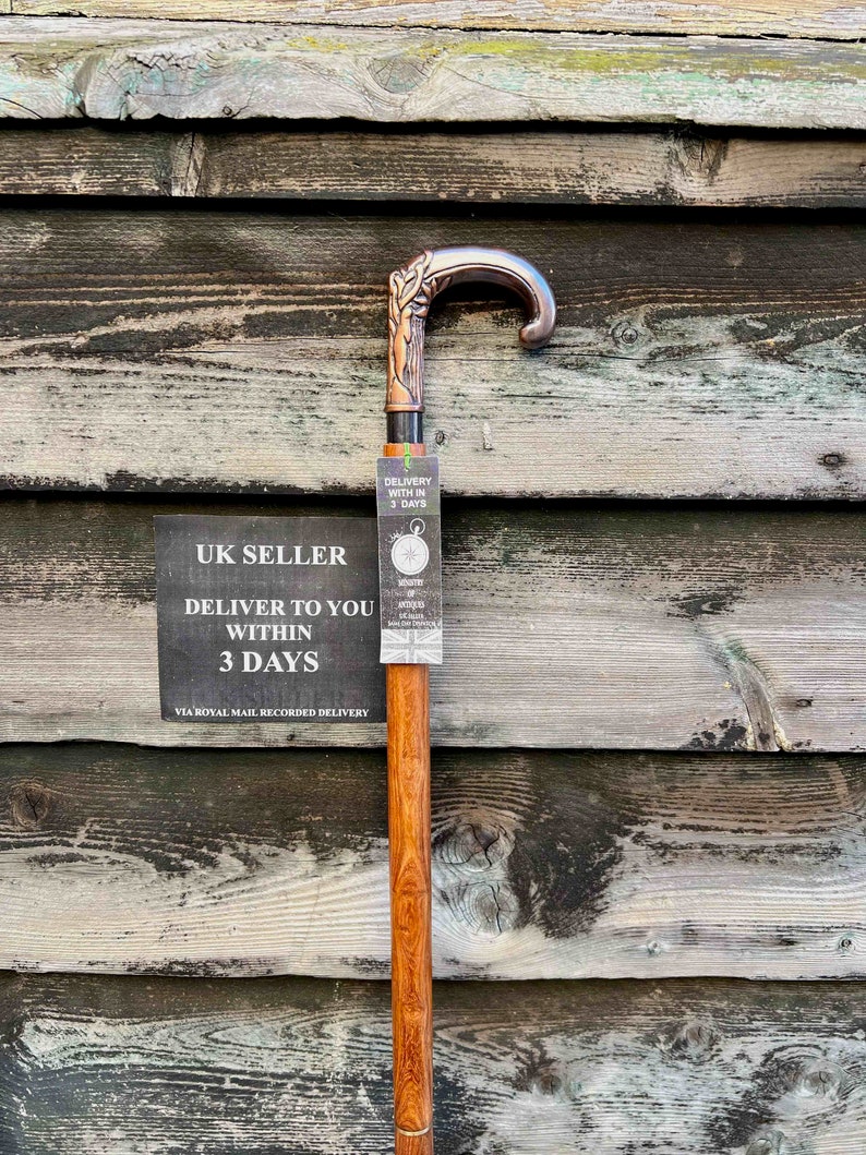 May include: A wooden walking stick with a copper-colored handle featuring a carved design. The stick is leaning against a wooden wall with a sign that reads "UK SELLER DELIVER TO YOU WITHIN 3 DAYS VIA ROYAL MAIL RECORDED DELIVERY".