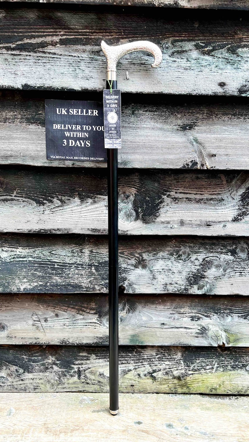 K&ouml;nnte beinhalten: Ein schwarzer Gehstock mit einem silbernen Griff. Der Griff hat ein dekoratives Design. Der Stock steht aufrecht an einer Holzwand.