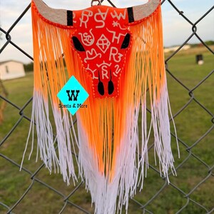 May include: An orange and white decorative bull skull with fringe, hanging from a brown string. The skull features a black nose and horns with a light brown color. The skull has white symbols and letters. The fringe transitions from orange to white.