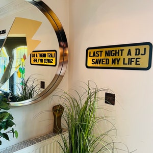 May include: A gold sign with black text that reads "Last Night A DJ Saved My Life" hanging on a white wall. A round mirror with a gold frame is reflected in the wall.