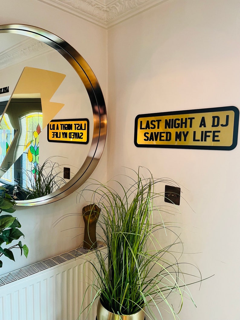 May include: A gold sign with black text that reads "Last Night A DJ Saved My Life" hanging on a white wall. A round mirror with a gold frame is reflected in the wall.
