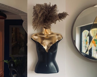 May include: A black mannequin torso with a gold painted bust and a dried pampas grass head. The mannequin is mounted on a white wall.