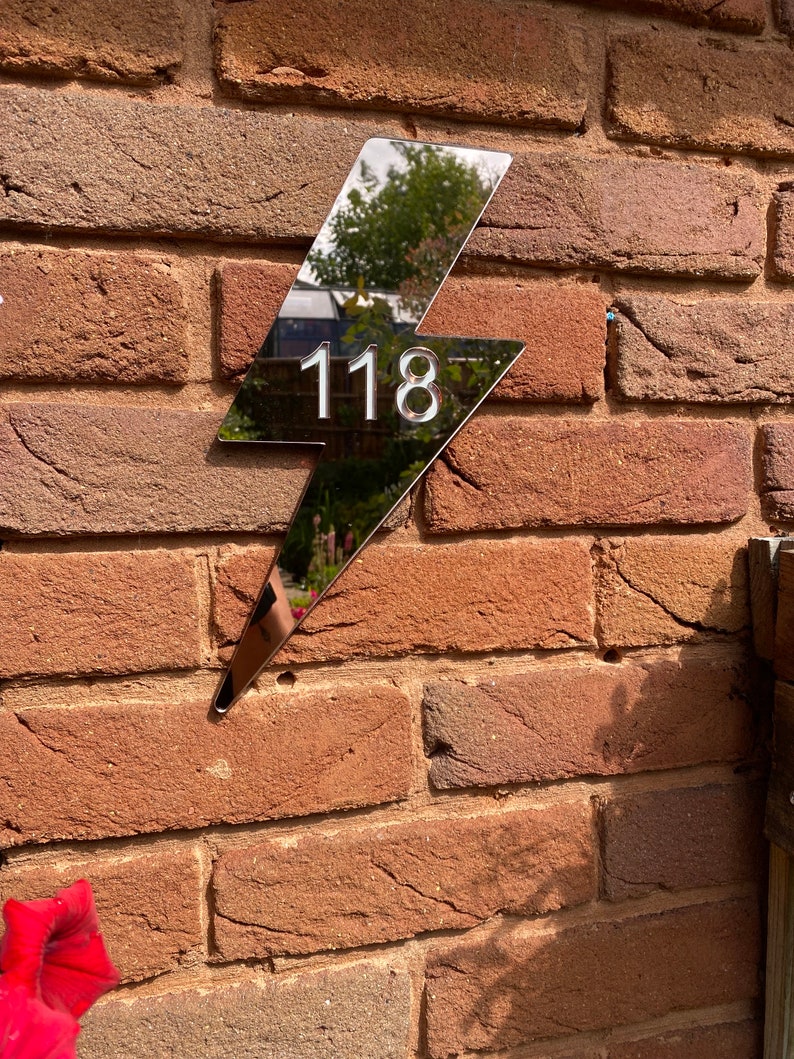 May include: A silver lightning bolt shaped house number sign with the number 118 on it. The sign is mounted on a brick wall.