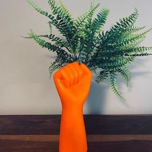 May include: A bright orange fist-shaped vase filled with green fern-like foliage. The vase is a bold orange colour and has a smooth finish. The plants are a vibrant green, providing a strong contrast against the vase.