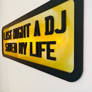 May include: A yellow sign with black lettering that reads "Last night a DJ saved my life". The sign is framed in black and has a glossy finish.