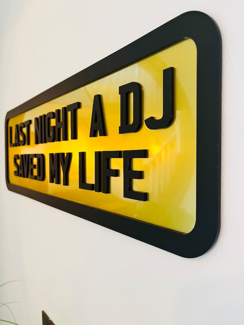 May include: A yellow sign with black lettering that reads "Last night a DJ saved my life". The sign is framed in black and has a glossy finish.