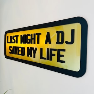 May include: A black framed sign with a yellow background and the text "Last night a DJ saved my life".