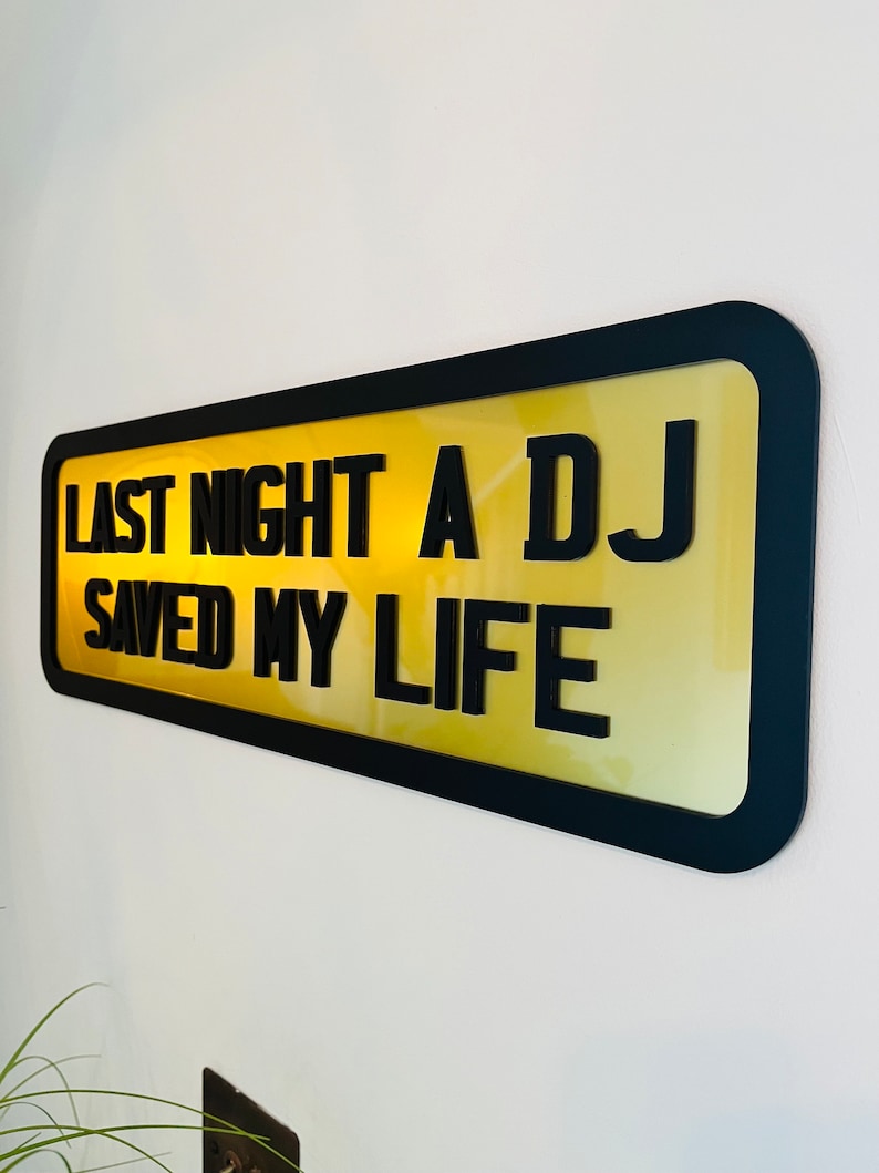 May include: A black framed sign with a yellow background and the text "Last night a DJ saved my life".