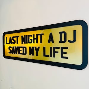 May include: A black framed sign with a yellow background and the text "Last night a DJ saved my life".