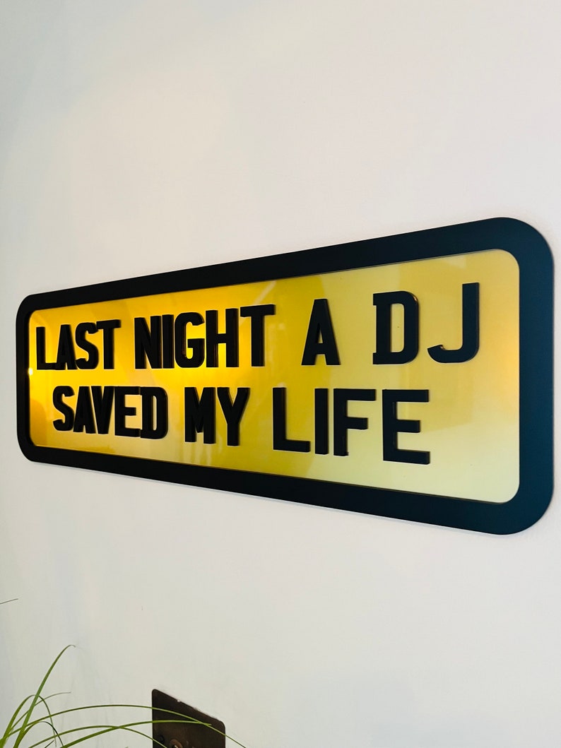 May include: A black framed sign with a yellow background and the text "Last night a DJ saved my life".