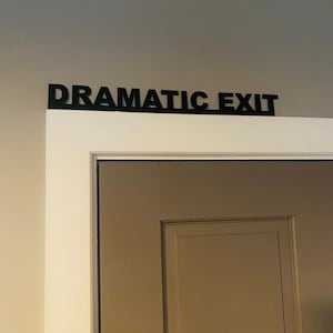 Puede incluir: Una puerta blanca con un letrero negro encima que dice "Salida Dram&aacute;tica".