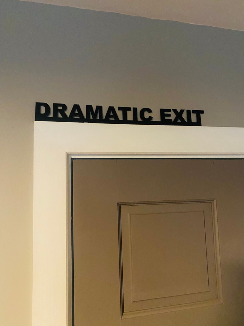 Puede incluir: Una puerta blanca con un letrero negro encima que dice "Salida Dram&aacute;tica".