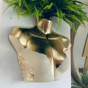 Può includere: Un busto in gesso dipinto d'oro con un motivo a stampa leopardata, utilizzato come vaso per una pianta di felce verde.