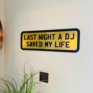 May include: Black and gold sign with the text "Last night a DJ saved my life".