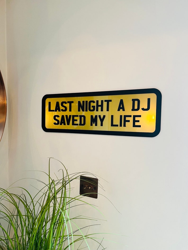 May include: Black and gold sign with the text "Last night a DJ saved my life".