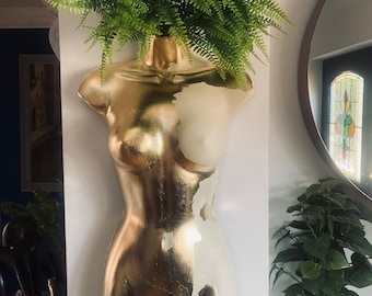 May include: A gold and white painted plaster mannequin torso with a green leafy plant growing out of the top of its head.