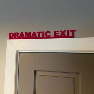 Puede incluir: Un marco de puerta blanco con un letrero rojo que dice "Dramatic Exit" sobre una puerta marr&oacute;n cerrada.