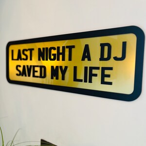 May include: A black framed sign with a gold background and the text "Last night a DJ saved my life".