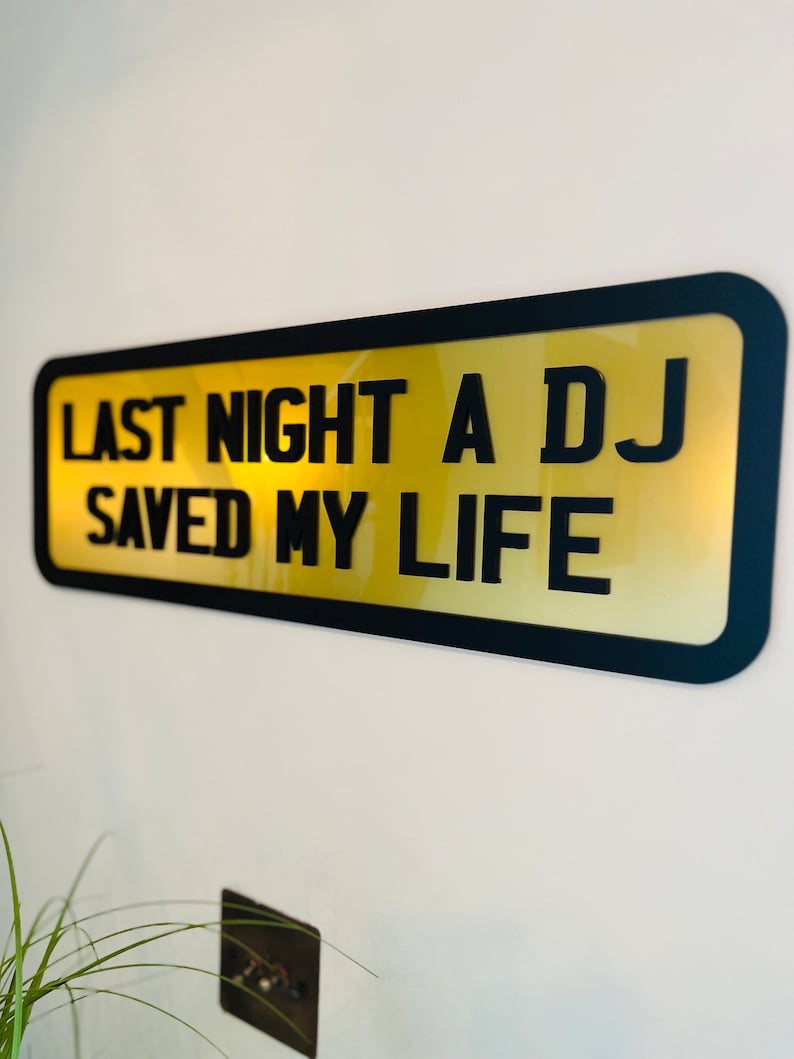May include: A black framed sign with a gold background and the text "Last night a DJ saved my life".