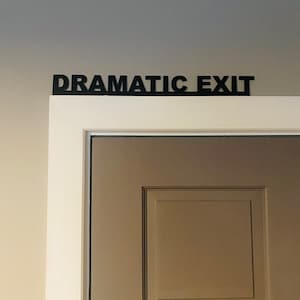 Puede incluir: Una puerta blanca con un letrero negro encima que dice "Salida Dram&aacute;tica".