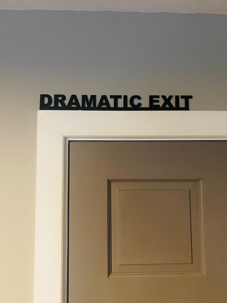 Puede incluir: Una puerta blanca con un letrero negro encima que dice "Salida Dram&aacute;tica".