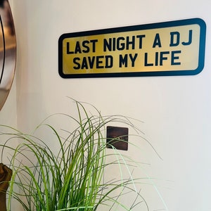 May include: A gold sign with black lettering that reads "Last Night A DJ Saved My Life" hanging on a white wall. A green plant is in the foreground.