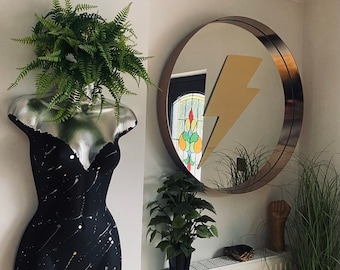 May include: A black and silver mannequin torso with a green plant growing out of the head. The mannequin is decorated with silver stars. A round mirror with a gold lightning bolt reflection is on the wall behind the mannequin.