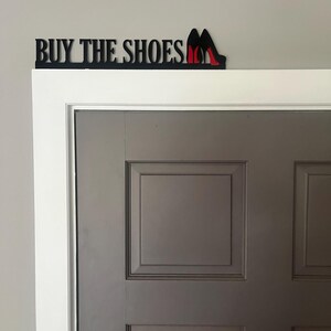 May include: Black metal sign with the words "BUY THE SHOES" and a pair of red high heels.