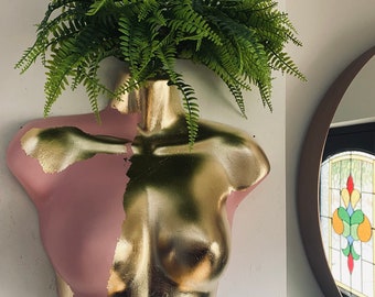 May include: A gold and pink torso planter with a green fern plant growing out of the top.