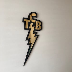 May include: A black and gold lightning bolt-shaped wall decor with the letters "TCB" in a stylized font. The letters and lightning bolt are gold, and the outline is black. The decor is mounted on a white wall.