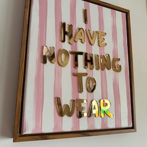 May include: Framed canvas art with a white and pink striped background. Gold metallic letters spell "I HAVE NOTHING TO WEAR". The last word has a rainbow-coloured "R". The frame is made of wood.