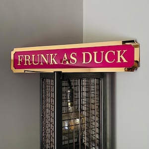 May include: A pink and gold sign with the text "FRUNK AS DUCK" on it. The sign is hanging from a black metal cage light fixture.