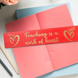May include: Red wooden bookmark with a gold inscription that reads "Teaching is a work of heart". The bookmark has two gold heart designs with leaves on either side of the text.