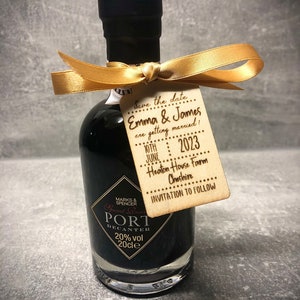May include: A small bottle of port wine with a gold ribbon and a wooden tag that says "Save the date Emma & James are getting married! 10th June 2023 Heaton House Farm Cheshire Invitation to follow"