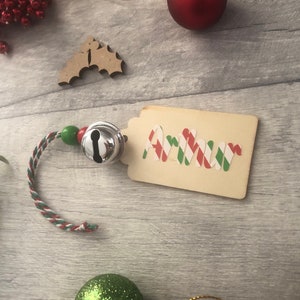 May include: A wooden gift tag with the name "Arthur" written in red, green, and white striped letters. The tag is attached to a red and green string with a silver bell.