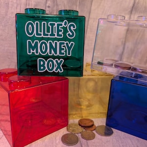 May include: Four clear plastic money boxes shaped like Lego bricks. The green money box has the text "Ollie's Money Box" on the front. The other money boxes are red, yellow, and blue.