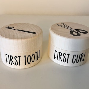 Puede incluir: Dos pequeñas cajas de madera con tapas. La caja de la izquierda tiene un icono de cepillo de dientes y el texto "FIRST TOOTH". La caja de la derecha tiene un icono de tijeras y el texto "FIRST CURL".