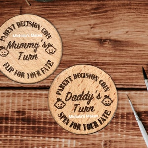 May include: Two wooden coin-shaped tokens with the text "PARENT DECISION COIN" on the top. One token says "Mummy's Turn" and the other says "Daddy's Turn". Both tokens have a baby face graphic and the text "Michala's Makes" on them.