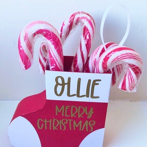 Puede incluir: Calcetín de Navidad rojo y blanco lleno de bastones de caramelo con el nombre "Ollie" y el texto "Feliz Navidad" en letras doradas.