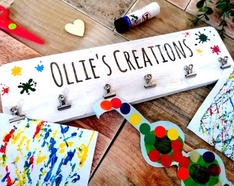 Könnte beinhalten: Ein weißes Holzschild mit dem Text "Ollie's Creations" und bunten Farbspritzern. Das Schild hat fünf Metallklammern zum Aufhängen von Kunstwerken. An den Klammern hängen drei bunte Kunstwerke.