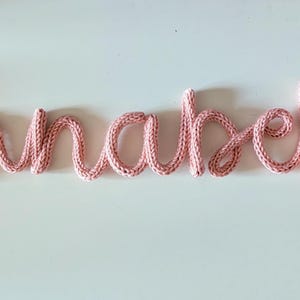 以下が含まれることがあります： 「Annabella」という名前を綴ったピンクのニット製のワードアート作品です。