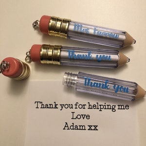 Puede incluir: Tres recipientes transparentes de plástico con forma de lápiz con tapas doradas y gomas de borrar rosas. Los recipientes tienen un mensaje escrito en el interior: "Mrs. Farrow", "Gracias", y "Gracias". Una tarjeta blanca con el mensaje "Gracias por ayudarme Amor Adam xx" está en primer plano.