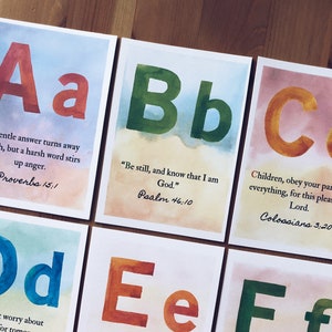 Könnte beinhalten: Ein Satz von 10 Aquarell-Alphabetkarten mit einem Bibelvers auf jeder Karte. Die Karten zeigen einen bunten Aquarellhintergrund mit einem großen Buchstaben in der Mitte. Die Buchstaben sind A, B, C, D, E, F, G, H, I und J. Die Verse stammen aus der Bibel und sind in schwarzer Schrift gedruckt.