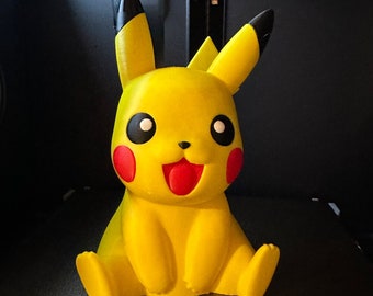 May include: A yellow Pikachu figurine with black ears and eyes, a red mouth, and red cheeks. The figurine is sitting on a dark surface.