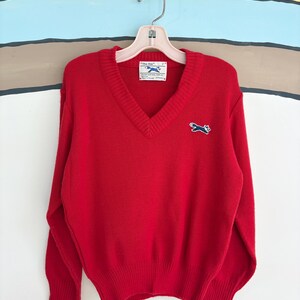 May include: A bright red V-neck sweater with long sleeves, hanging on a hanger. The sweater features a small embroidered fox logo on the chest and a tag that reads "The Fox". The sweater is made of a soft knit material.