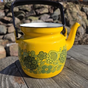 Puede incluir: Una tetera vintage de esmalte amarillo con asa y boquilla negras. La tetera presenta un diseño floral en tonos verdes. El asa es negra y el interior es blanco. Un encantador artículo de cocina.